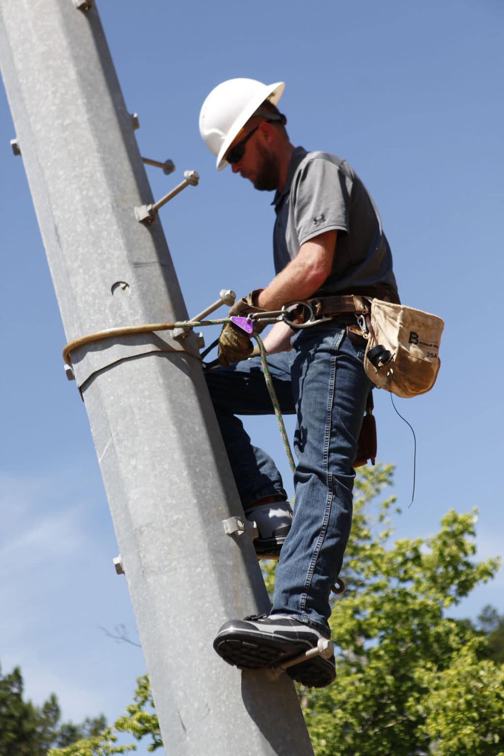 A worker in jeans, a gray shirt, and a hard hat is climbing a metal pole secured with a harness. Wearing FRO4 Series Quatro Dielectric 2 Buckle Overshoes and gloves, he has a tool pouch around his waist. The sky is clear and blue with trees in the background.