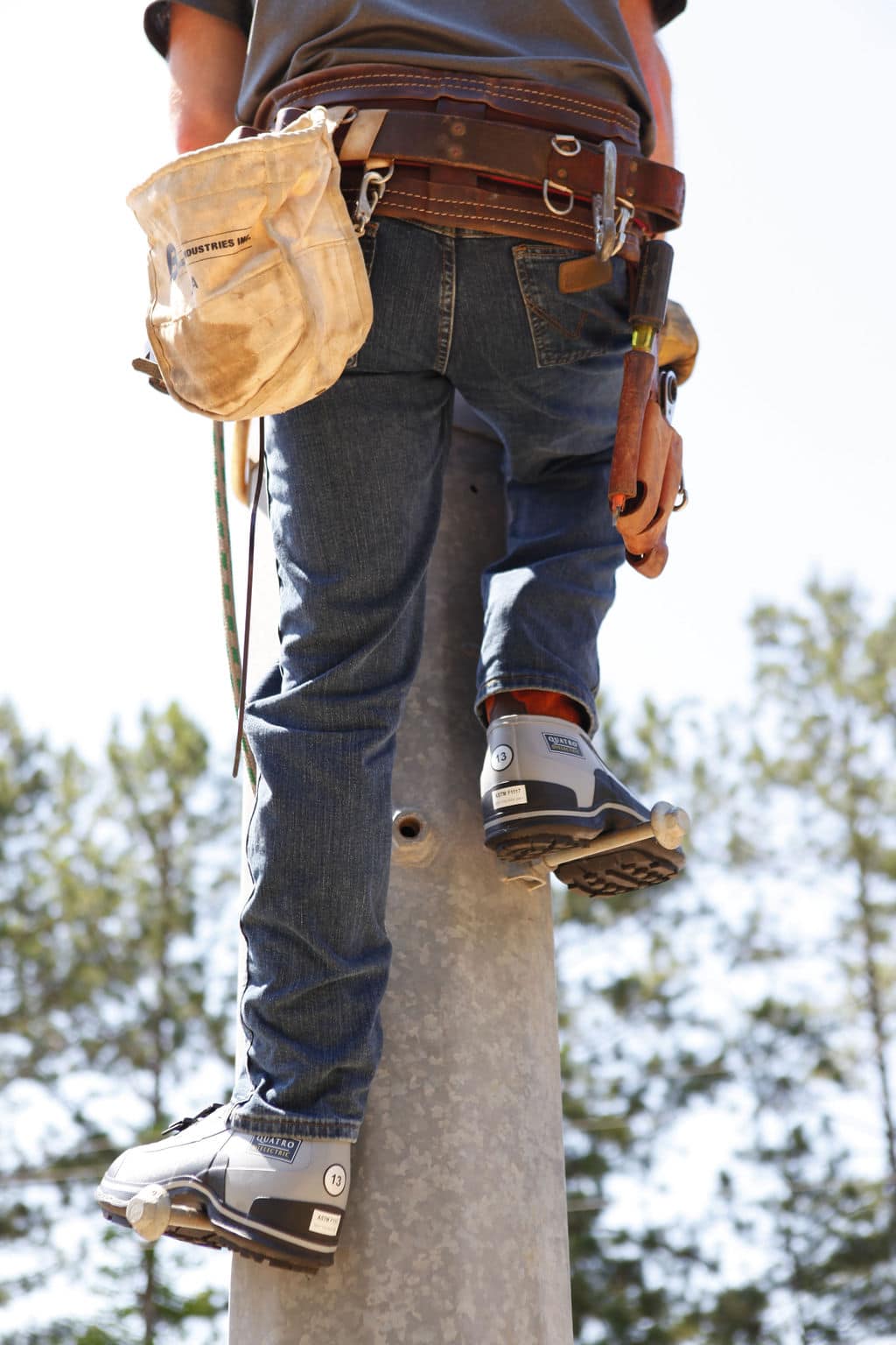 A person climbing a pole while wearing a tool belt, jeans, and climbing boots with FRO4 Series Quatro Dielectric 2 Buckle Overshoes. They are using safety gear and standing against a clear sky with trees in the background.