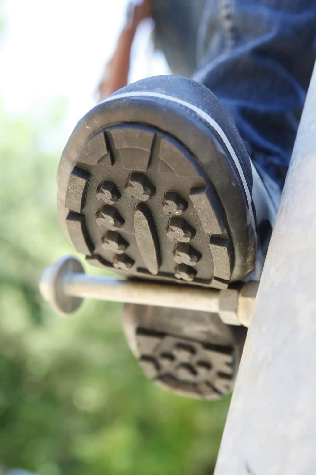 Close-up of a rugged boot sole gripping a metallic step, focusing on the tread pattern. The background is blurred, showing green foliage. It looks like the FRO3 Series Quatro Dielectric Slip On Overshoes are worn by someone climbing or standing on an outdoor structure.