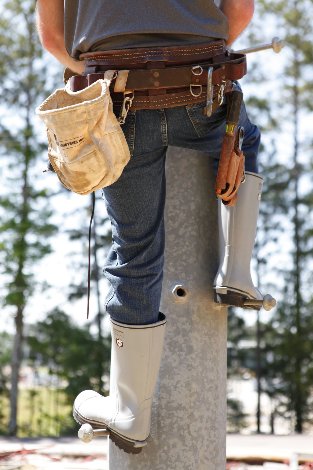 A person in blue jeans and rugged FQD9 Series Quatro Dielectric Thermal Boots is climbing a utility pole. Equipped with a tool belt and a white pouch brimming with various tools, they work efficiently against a backdrop of green trees and bright sky.