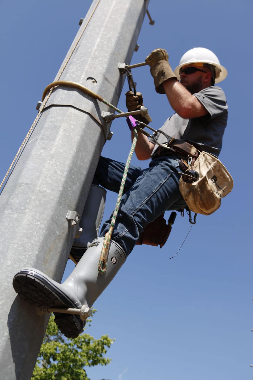 Under a clear blue sky, a utility worker in a hard hat and safety gear climbs a pole using a harness and safety equipment. Equipped with tools, a bag, and FQD9 Series Quatro Dielectric Thermal Boots, the worker ensures durability and protection during the climb.