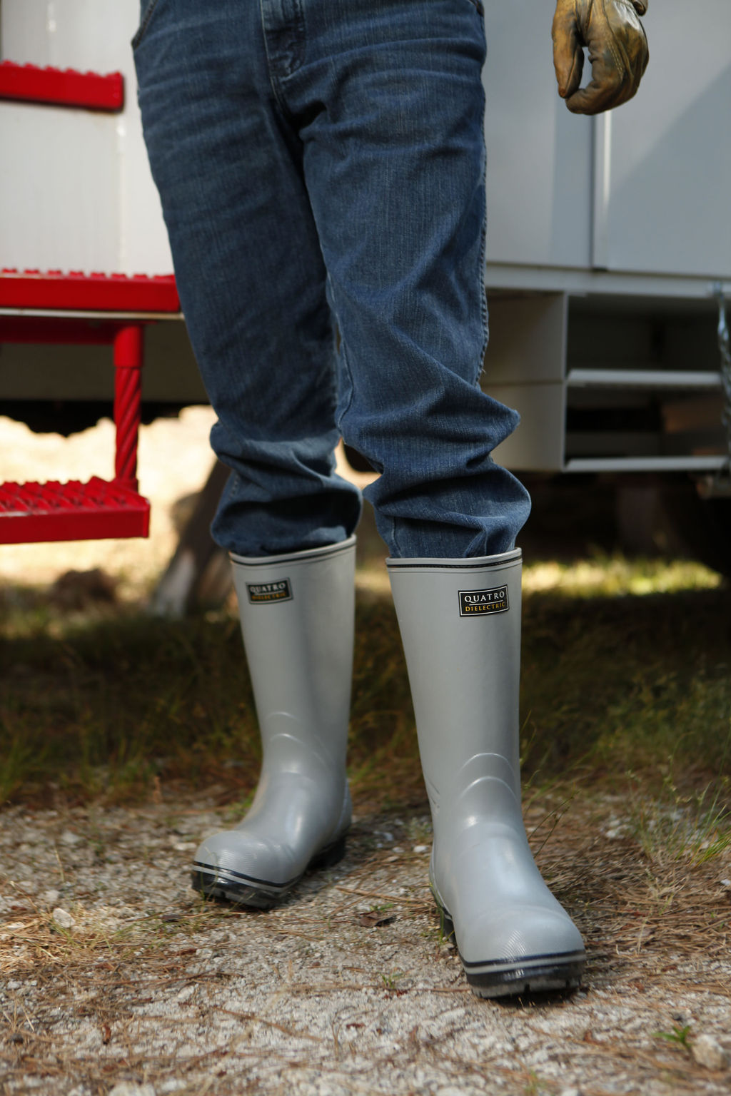 A person wearing blue jeans and gray FQD9 Series Quatro Dielectric Thermal Boots stands beside a vehicle with red steps. One hand is wearing a brown glove and rests by their side, while grass and gravel cover the ground.