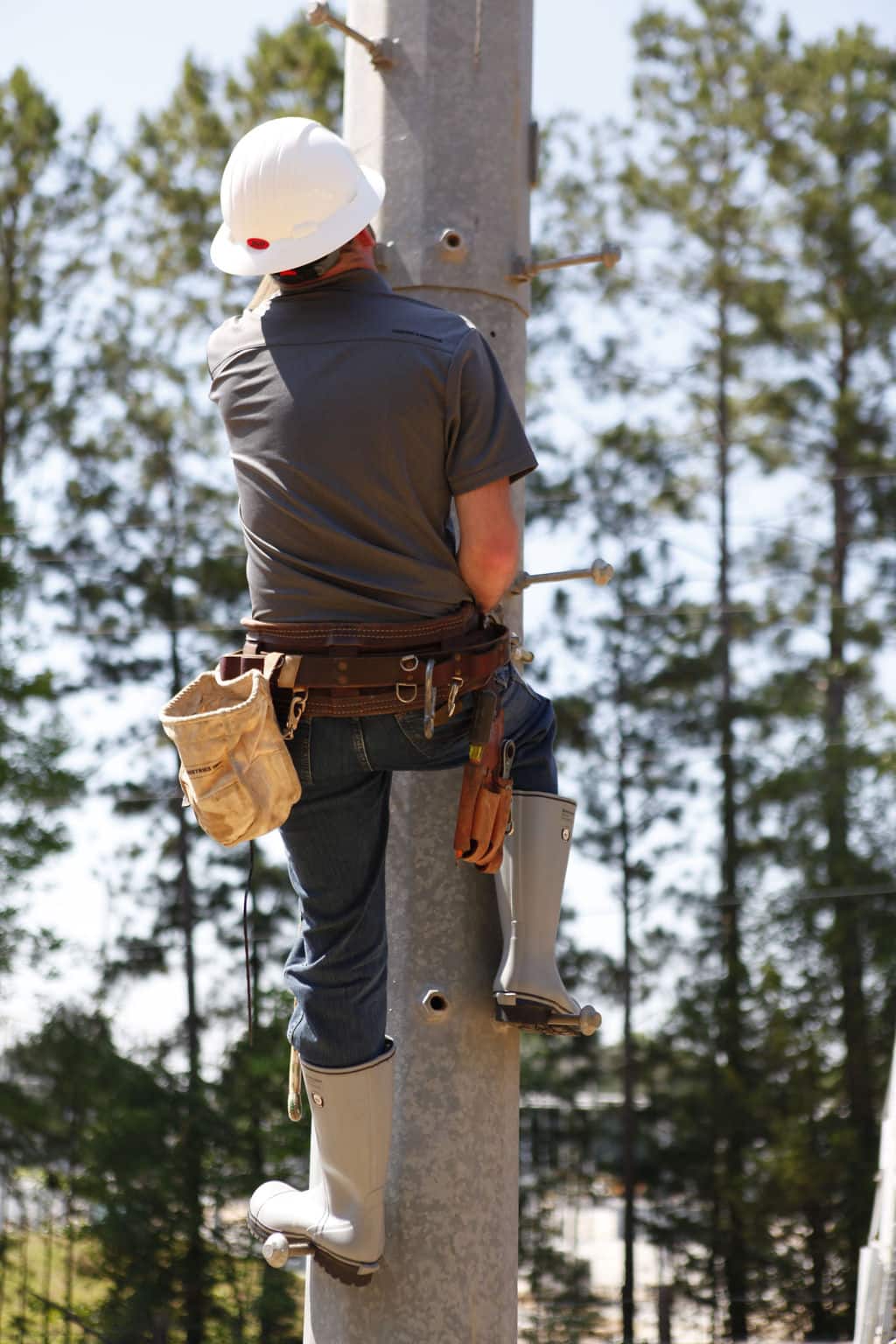 A worker in a hard hat and safety gear climbs a utility pole, confidently tackling heights in FQD9 Series Quatro Dielectric Thermal Boots. The person dons a tool belt amid a forested backdrop, ensuring safety and style with every step.